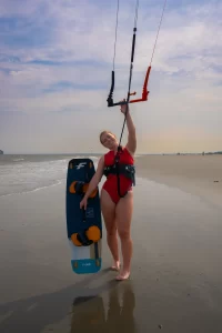 Girl in red one piece holds up blue F-One kiteboard and kite on the beach
