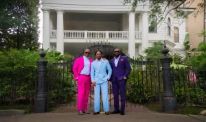 Three men in bright colored suites: blue, pink, and purple standing in front of a gate with the restraunt Columns in the back in New Orleans